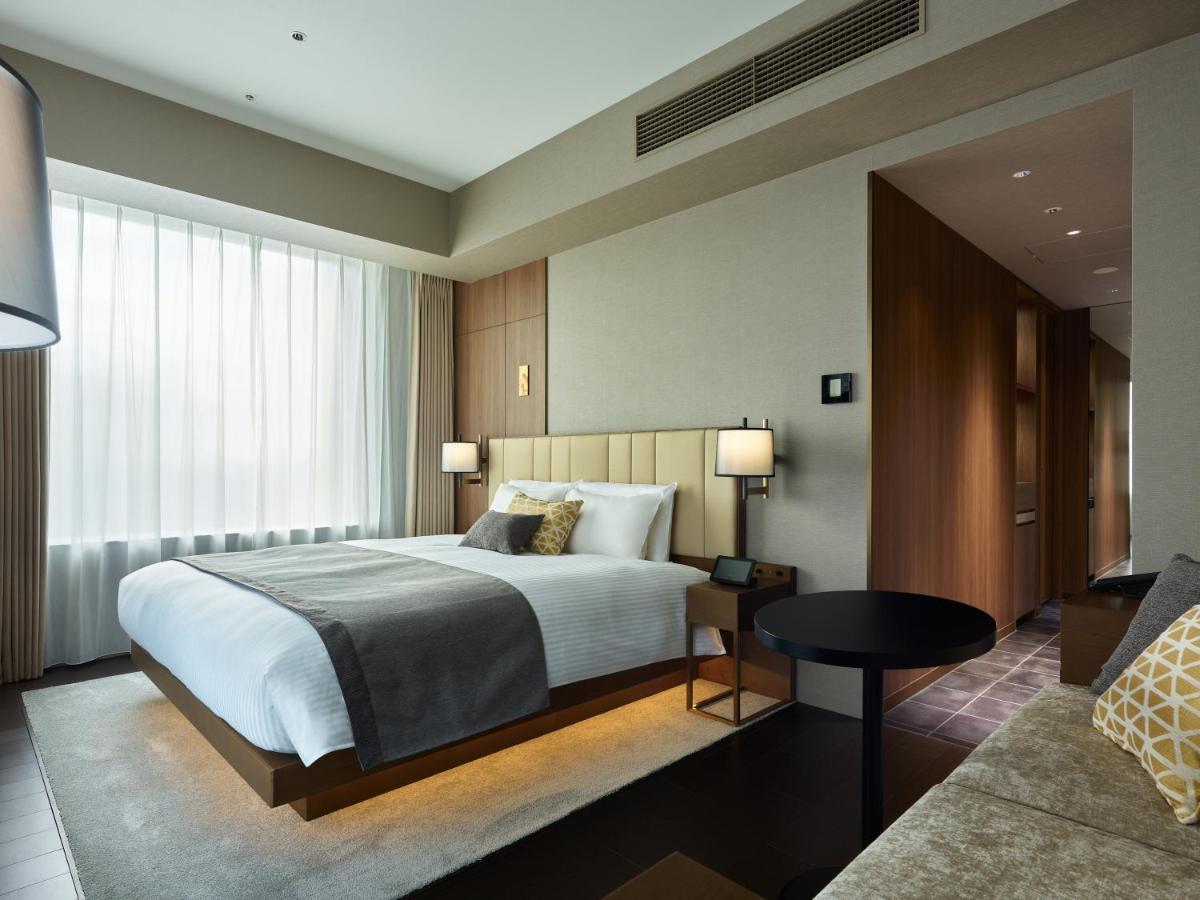 Mitsui Garden Hotel Roppongi Tokyo Premier Rom bilde The photo shows a modern hotel room. It features a large bed with a light-colored duvet and decorative pillows. There are bedside tables with lamps on either side of the bed. The room has large windows dressed with sheer curtains, allowing natural li