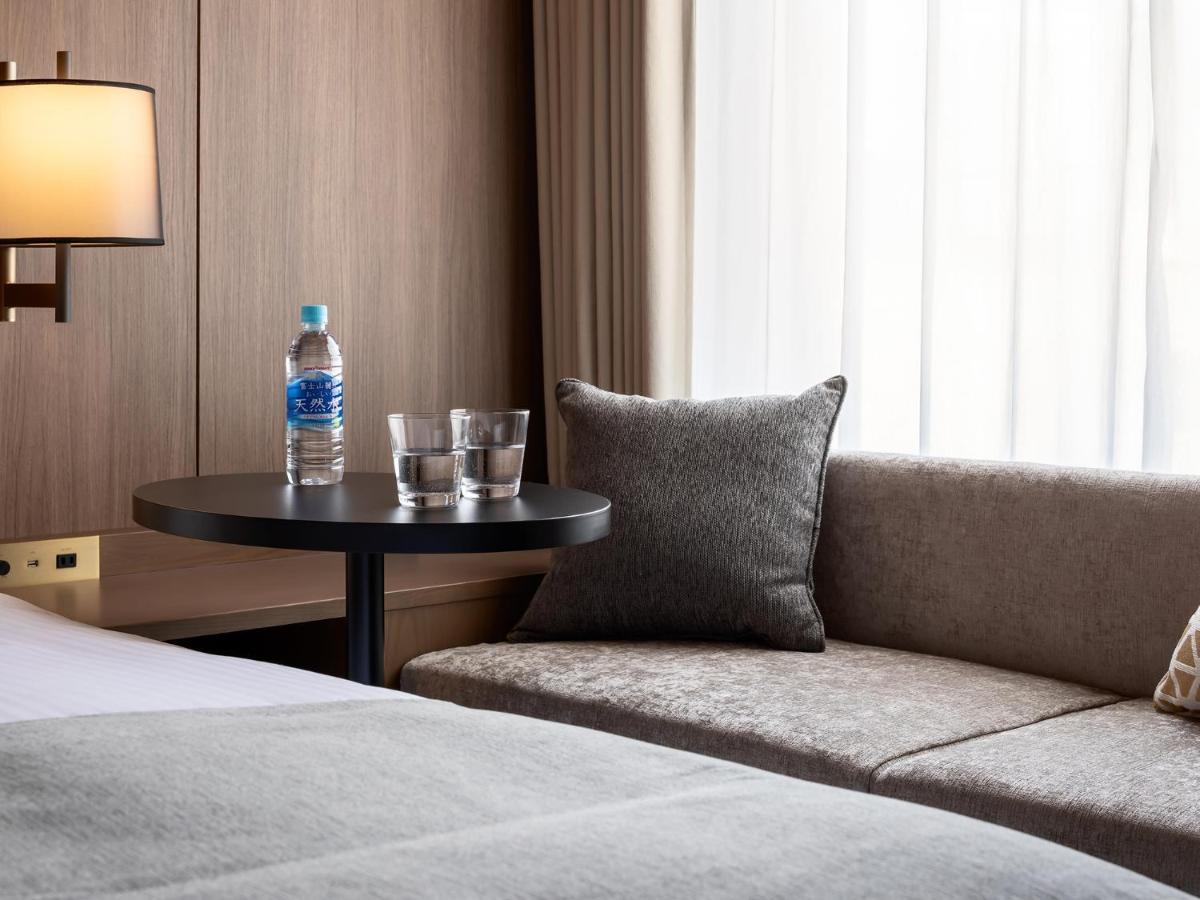 Mitsui Garden Hotel Roppongi Tokyo Premier Eksteriør bilde The photo depicts a cozy hotel room setting. On the left, there is a bedside table with a lamp. Against the wall, there is a comfortable-looking couch with a decorative pillow. In front of the couch, there's a small round table that holds a bottle of