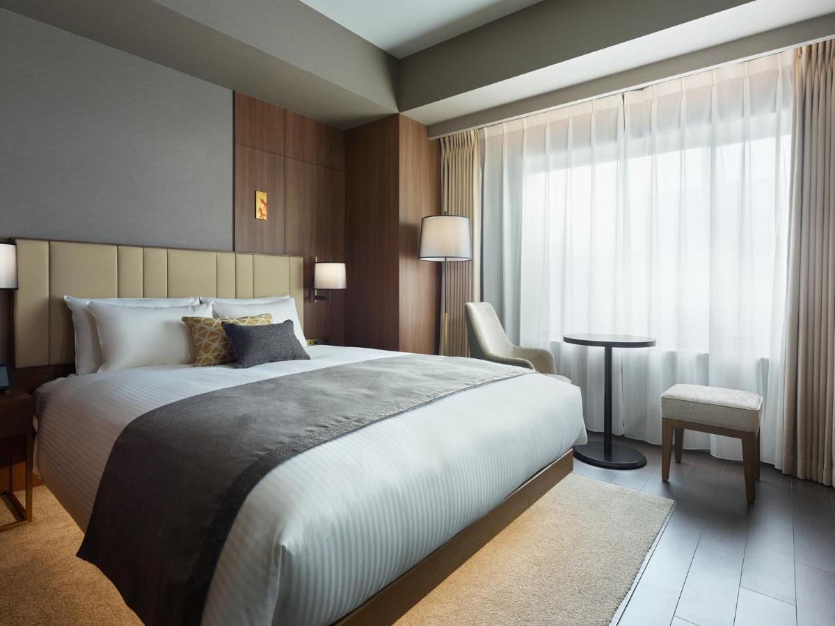 Mitsui Garden Hotel Roppongi Tokyo Premier Eksteriør bilde The photo shows a modern hotel room featuring a comfortable bed with white linens and a decorative throw pillow. The room has a neutral color palette with wooden accents on the walls. There are two bedside lamps and a chair positioned next to a small