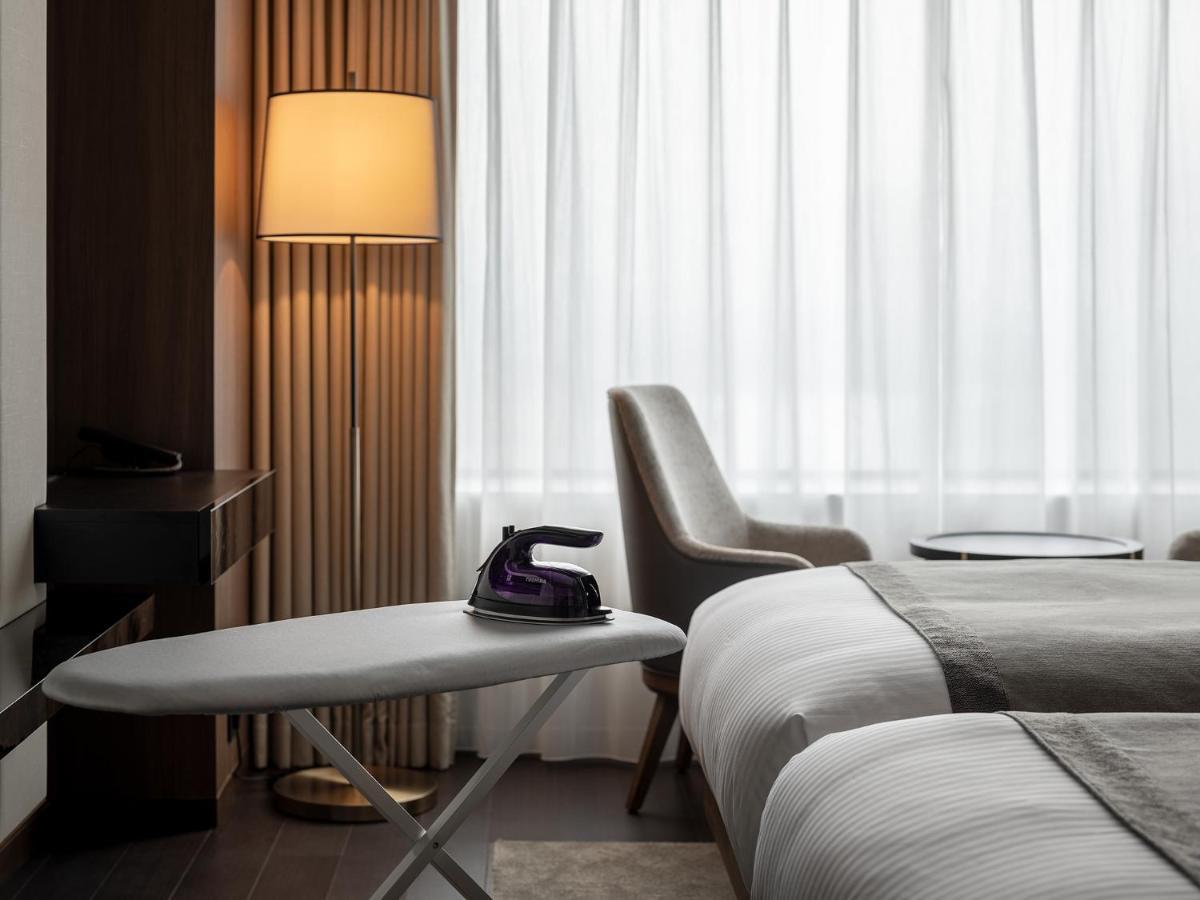Mitsui Garden Hotel Roppongi Tokyo Premier Eksteriør bilde The photo shows a modern hotel room featuring two beds with white bedding. There is a light-colored armchair next to a small side table. On the table, there is an iron, and beside it, a lamp with a warm glow is illuminating the space. The large windo