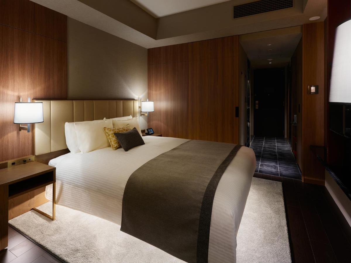 Mitsui Garden Hotel Roppongi Tokyo Premier Eksteriør bilde The image shows a modern hotel room with a warm and elegant design. In the center, there is a neatly made bed with white linens and a dark gray throw blanket. Flanking the bed are two bedside lamps providing soft lighting. The walls have a rich wood 