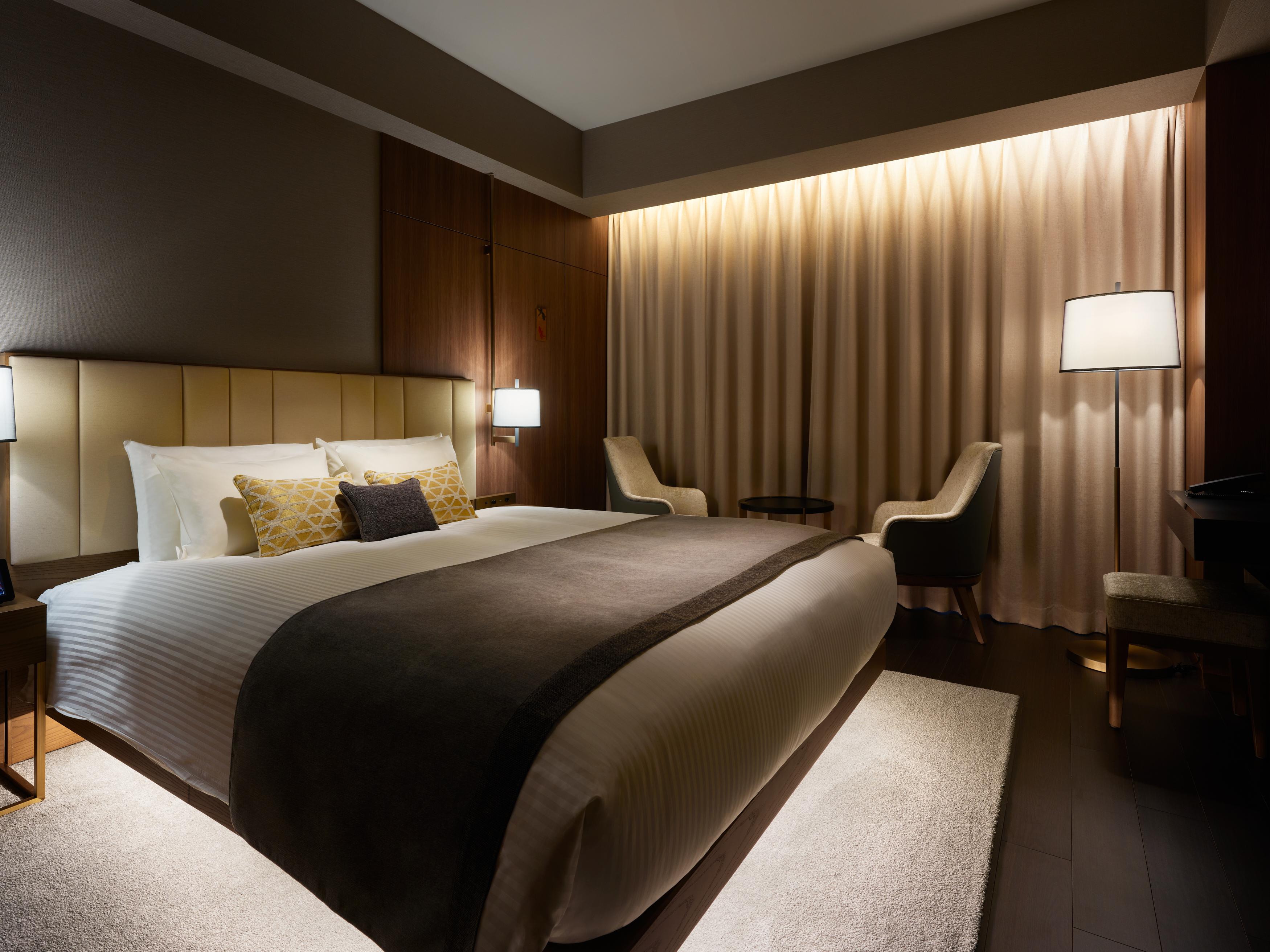 Mitsui Garden Hotel Roppongi Tokyo Premier Eksteriør bilde The photo depicts a modern hotel bedroom. It features a large bed with white linens and a dark gray throw blanket, accented by decorative pillows. The room has a warm ambiance created by soft lighting, including two bedside lamps and a lighted area b