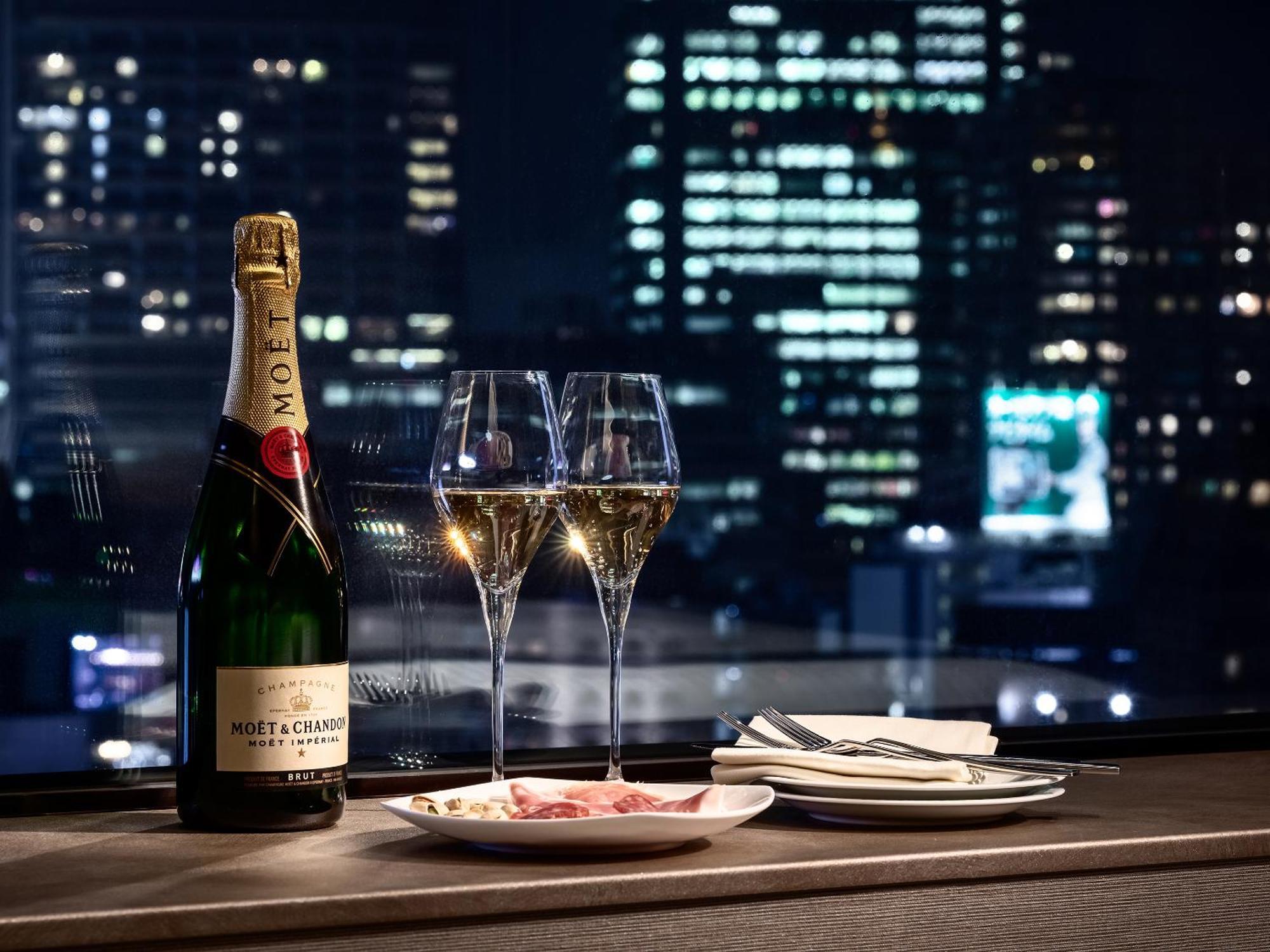 Mitsui Garden Hotel Roppongi Tokyo Premier Eksteriør bilde The photo features a bottle of champagne, specifically Moët & Chandon, alongside two filled champagne glasses. The table setting includes a small plate with a selection of food and neatly arranged utensils. In the background, there is a view of a cit