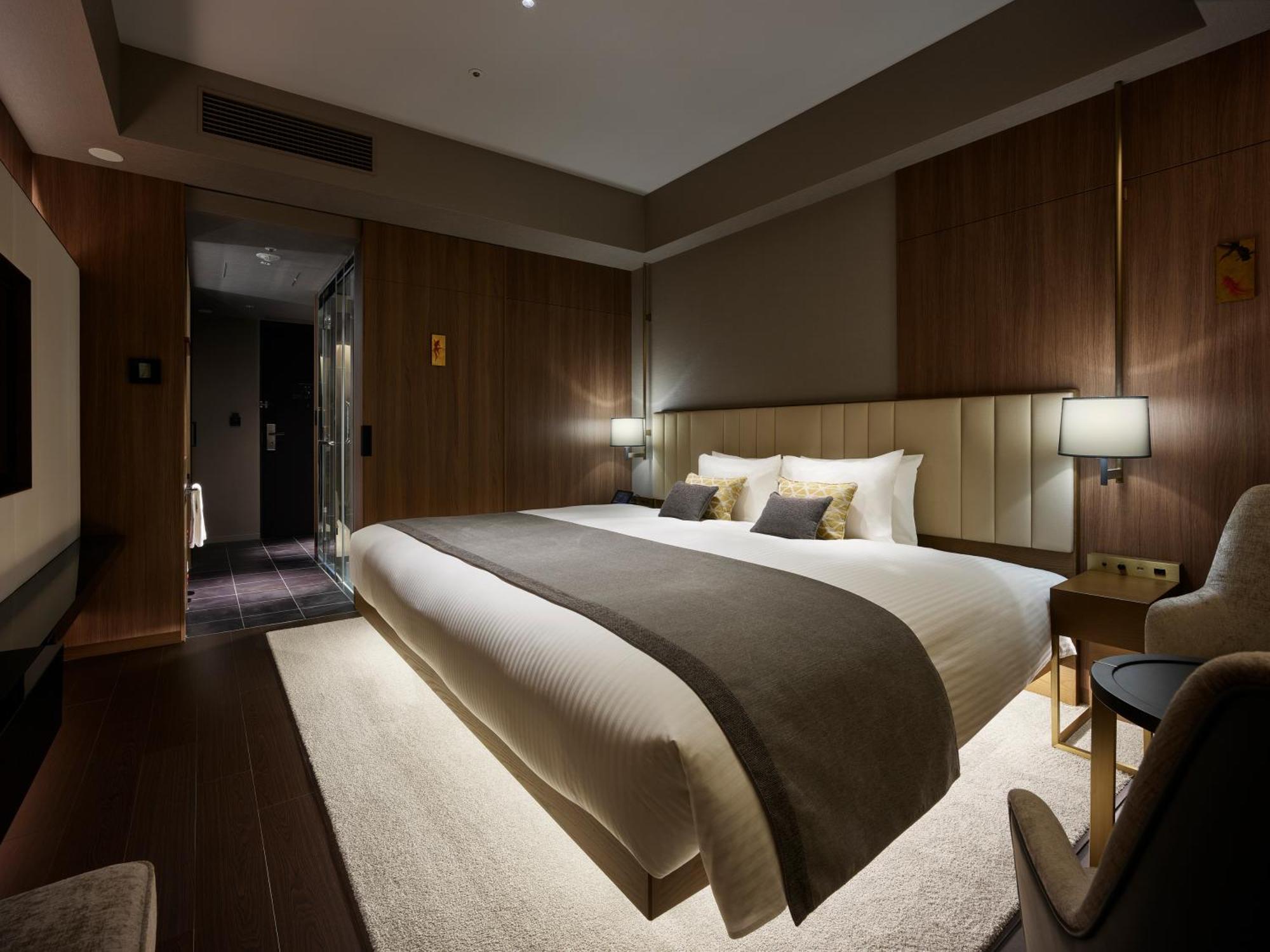 Mitsui Garden Hotel Roppongi Tokyo Premier Eksteriør bilde The photo shows a modern hotel room featuring a large bed with a neutral-colored duvet and a dark throw blanket. The headboard is upholstered, and there are two bedside lamps on either side of the bed. The walls have a warm wood paneling, contributin