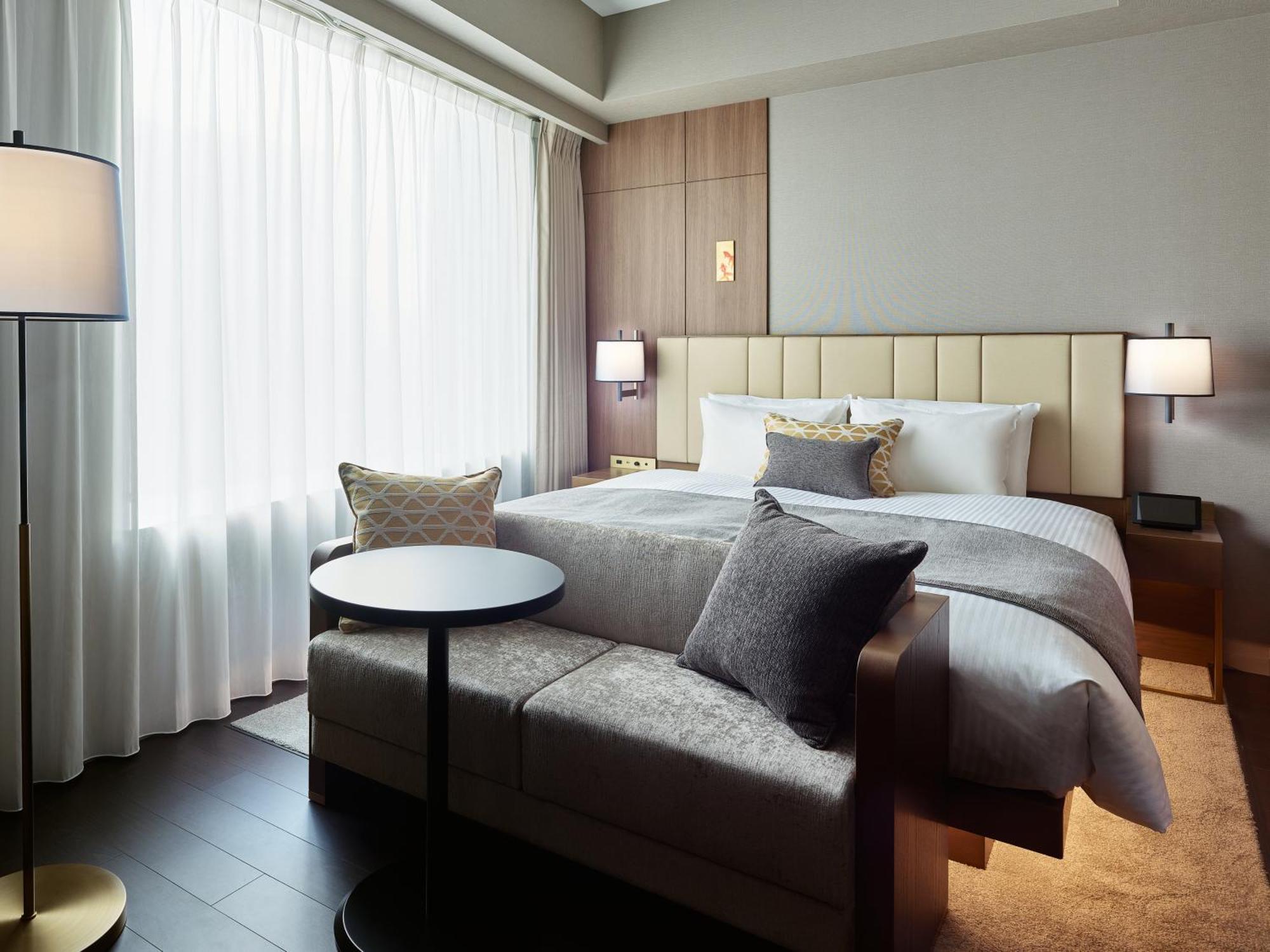 Mitsui Garden Hotel Roppongi Tokyo Premier Eksteriør bilde The photo shows a modern hotel room that features a large bed with a padded headboard and soft bedding. There's a small, round table accompanied by a couch or seating area, adorned with decorative pillows. To the left, a floor lamp provides additiona