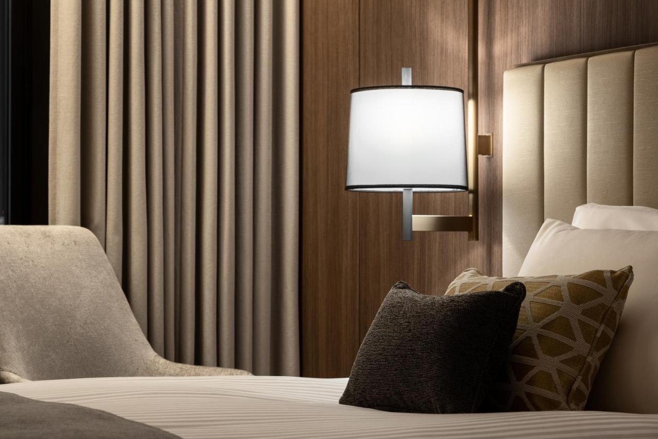 Mitsui Garden Hotel Roppongi Tokyo Premier Eksteriør bilde The photo shows a cozy hotel bedroom setting. There is a neatly made bed with white linens and a few decorative pillows, including a dark one and another with a geometric pattern. To the side, there is a soft armchair and a modern wall lamp mounted a